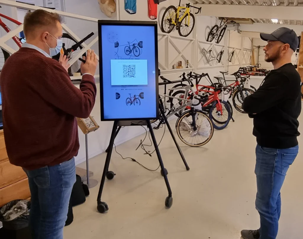 To mænd i en sort og rød trøje står foran et inaktiv whiteboard i en cykelbutik