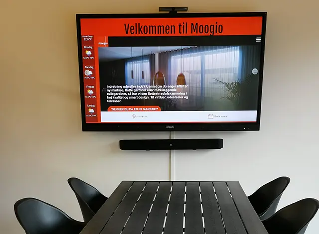 En skærm med et billed og skriven Velkommen til Moogio hænger på en hvid væg, under står et sort bord med 4 stole
