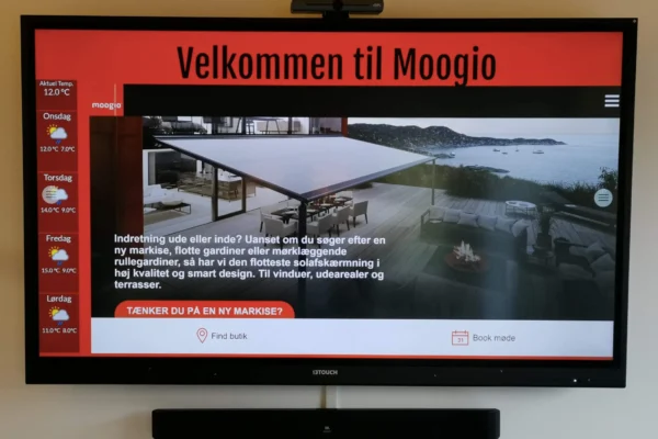 En skærm med et billed og skriven Velkommen til Moogio hænger på en hvid væg