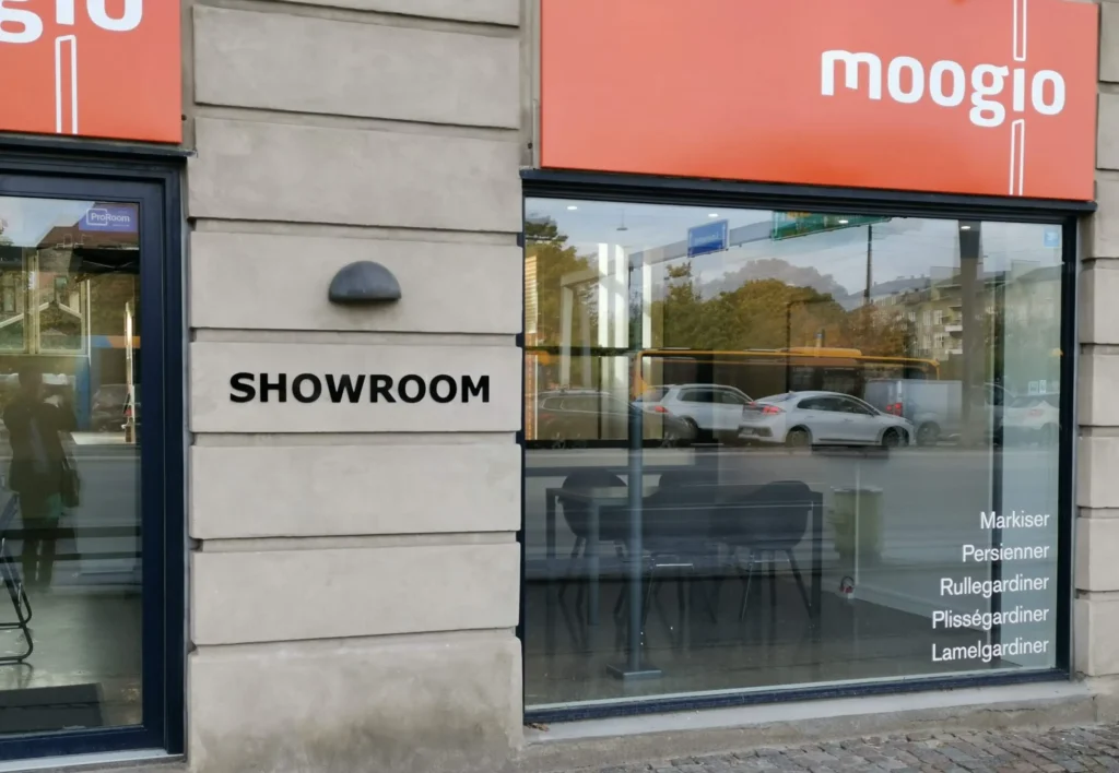 Et billede taget moogio showroom taget udefra. Der er et stort vindue med moogio orange logo for oven